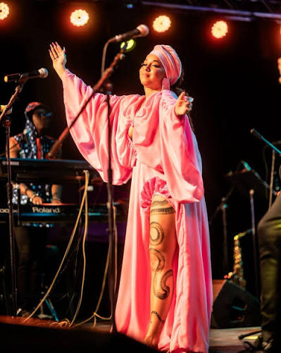 Riva performing in a flowing pink gown with painted serpent motif under stage lights